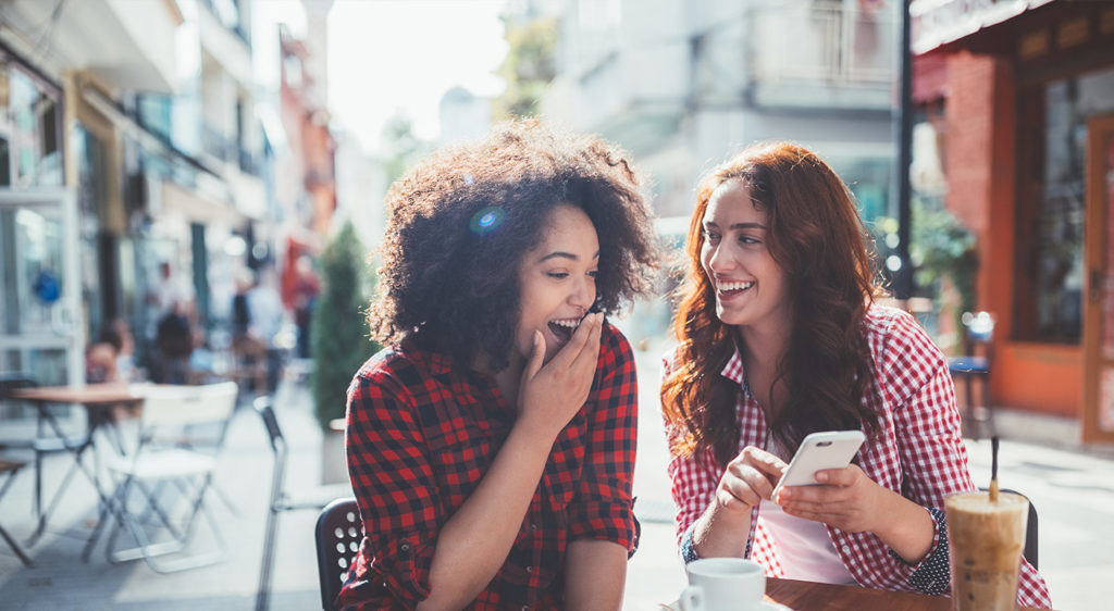 Two women in red flannel shirts sit outside a local cafe and laugh at content on a smartphone while drinking coffee.