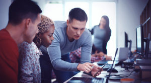 A man in a grey sweater points to his smart phone as he shows something to two colleagues in an office setting.